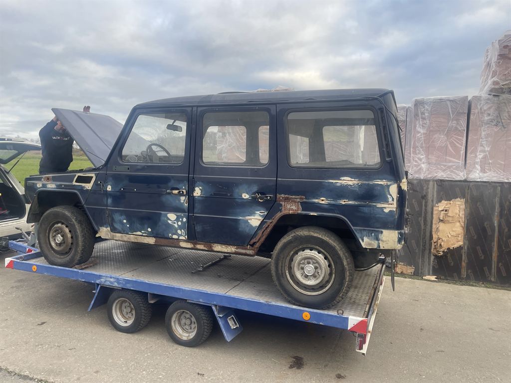 Anlieferung: der dunkelblaue G steht mit offener Motorhaube auf einem blauen Autoanhänger, an den Schwellern klafft der Rost.